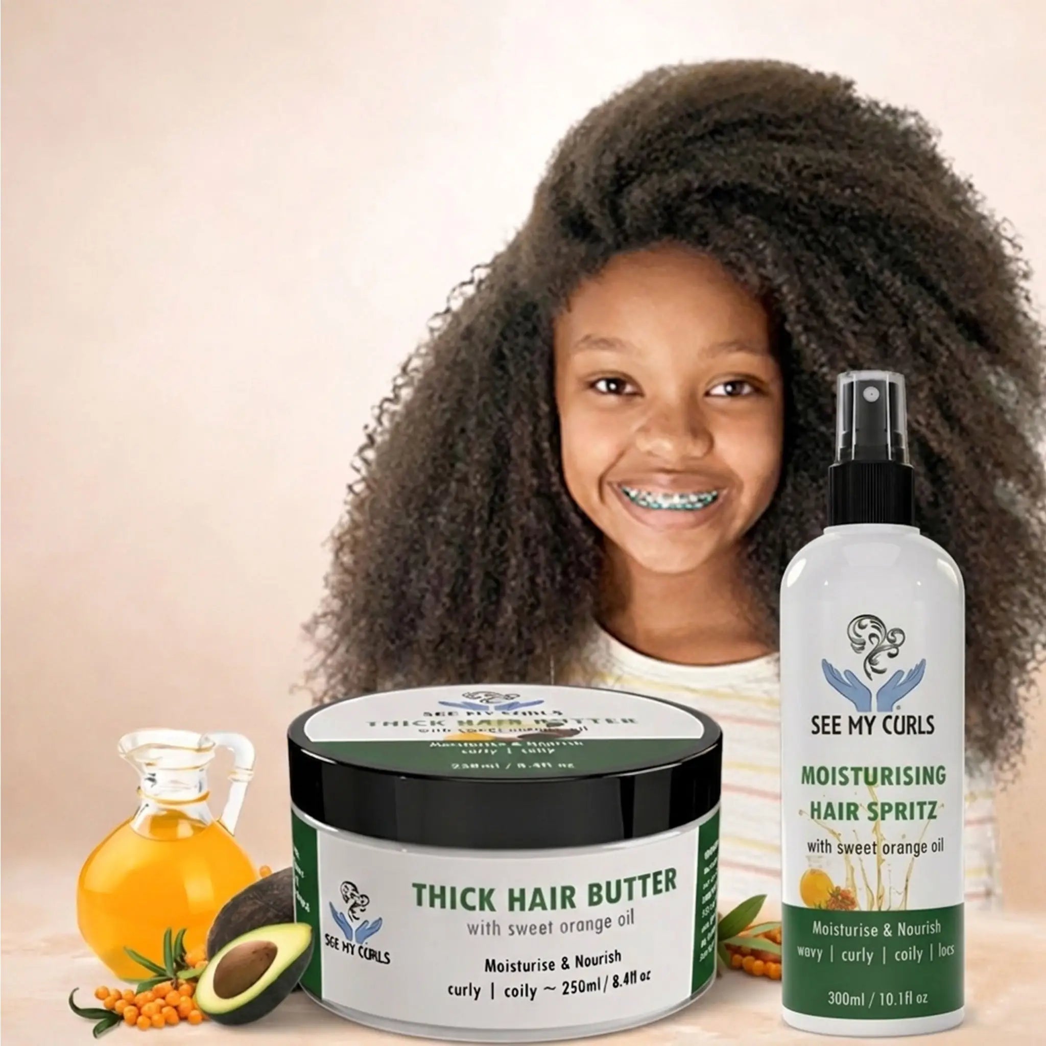 A young girl with a large, beautiful afro and voluminous coily hair smiling next to See My Curls "Thick Hair Butter" and "Moisturising Hair Spritz." The products feature sweet orange oil and are shown alongside natural ingredients like fresh avocado and a bottle of essential oil to highlight deep moisture for coily and kinky hair textures.