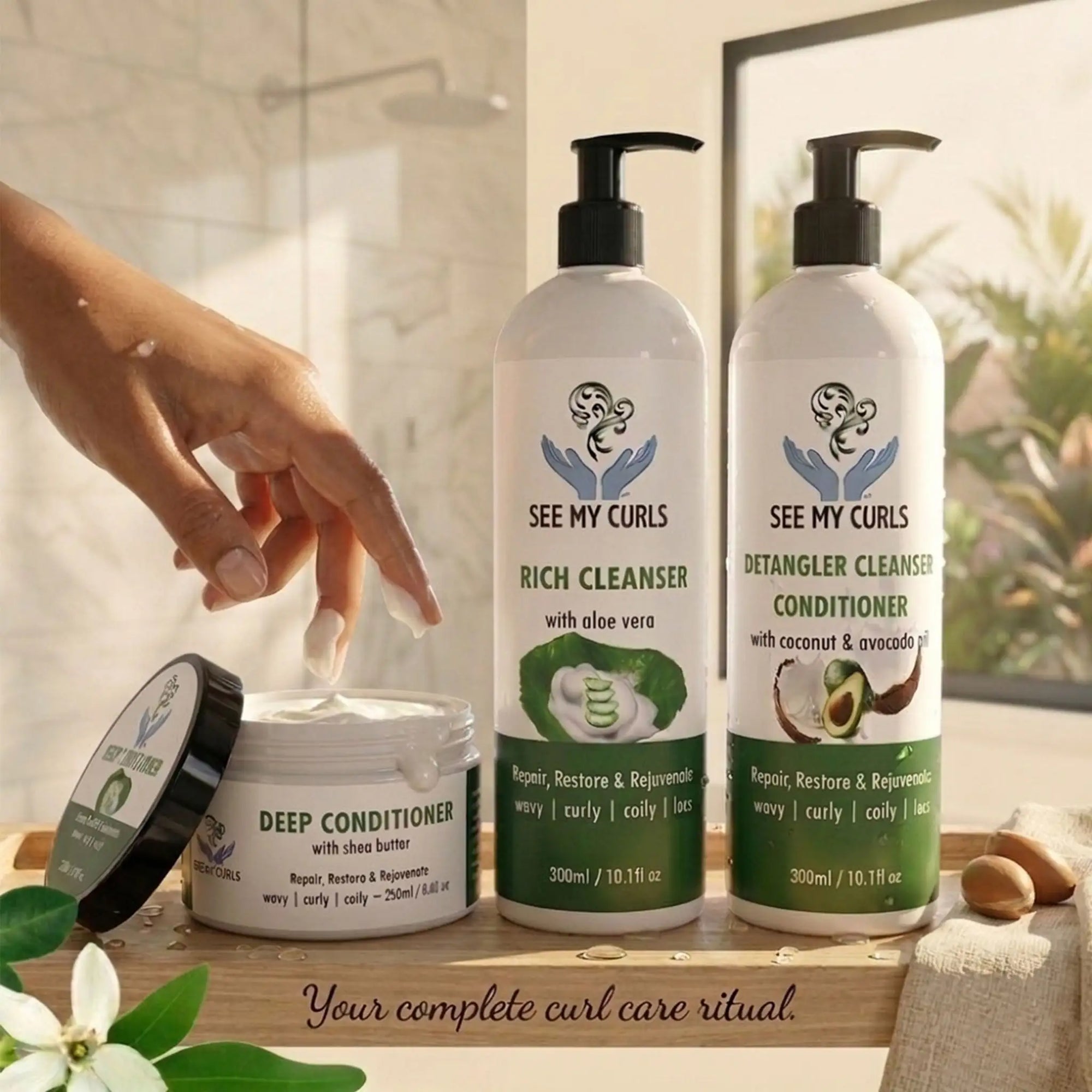A lifestyle shot of the See My Curls hair care line on a wooden tray in a bright bathroom. From left to right: a 250ml Deep Conditioner jar with its black lid resting nearby, a 300ml Rich Cleanser pump bottle (aloe vera), and a 300ml Detangler Cleanser Conditioner pump bottle (coconut & avocado). A hand is shown dipping fingers into the thick white deep conditioner.